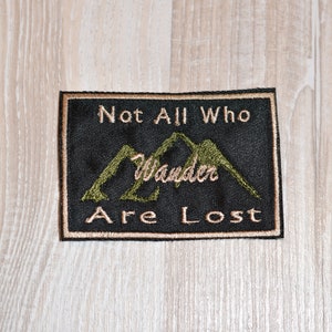 May include: Embroidered patch with the text "Not All Who Wander Are Lost" in gold thread on a black background. The patch features a mountain range design in green thread and a gold border.