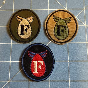 May include: Three embroidered patches with a bomb and wing design, each featuring the letter "F". One patch is on a dark green background, another on tan, and the third on black. The patches are circular with a contrasting border.