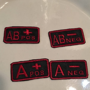 May include: Four black fabric patches with red embroidered lettering and symbols. The patches indicate blood types: AB+, AB-, A+, and A-.