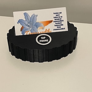 May include: A black circular stand with a ridged exterior and a central slot holding a white business card. The card features a blue lily illustration, the text "Tennesseelily", and social media handles. A circular button labeled "TAP PHONE" is centered on the front.