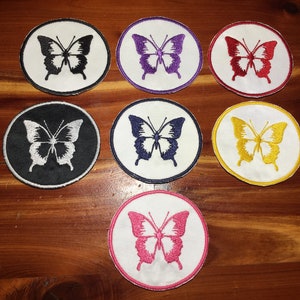 May include: Seven embroidered iron-on patches featuring a white butterfly with different colored outlines. The outlines are black, purple, red, yellow, navy blue, pink, and gold.