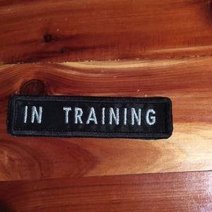 May include: Black rectangular patch with white embroidered text that reads "IN TRAINING".
