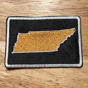 May include: A black and white embroidered patch featuring the outline of the state of Tennessee in gold.