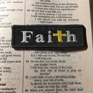 May include: A black fabric patch with white embroidered text that reads "Faith" and a yellow cross in the center.