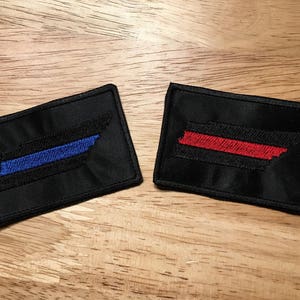 May include: Two black fabric patches in the shape of the state of Tennessee. One patch has a blue stripe across the state, and the other has a red stripe across the state.