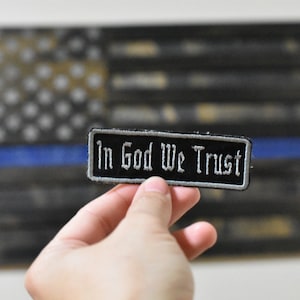 May include: A black and silver embroidered patch with the text "In God We Trust" on a black background. The patch is being held in a hand in front of a blurred image of an American flag.