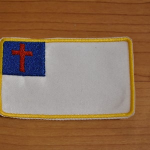 May include: A white patch with a yellow border and a blue and red embroidered cross. The cross is red with a white outline and is centered on the blue section of the patch.