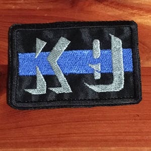 May include: Black embroidered patch with a blue stripe and a white stylized letter "K" and "D" design.