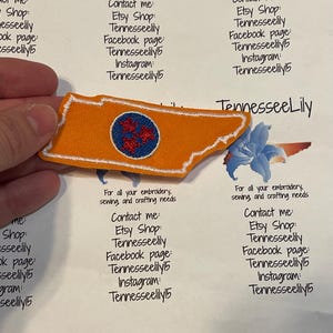 May include: An orange embroidered patch in the shape of Tennessee, with a white border. The patch features a blue circle with a red floral design. The text "TennesseeLily" is also visible.