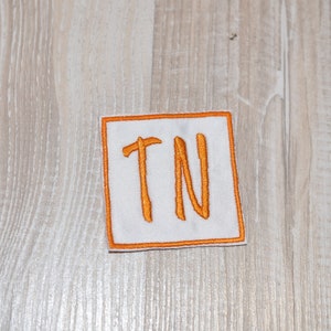 May include: A white square patch with an orange embroidered "TN" inside a thin orange border.
