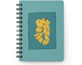 Puede incluir: Una libreta azul verdosa con encuadernación en espiral. La portada presenta una ilustración simple de un racimo de plátanos amarillos colgando de un gancho dentro de un rectángulo azul verdoso.
