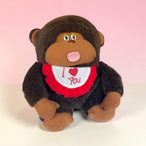 May include: A plush monkey with brown fur, tan hands and feet, and a brown face. It wears a red and white bib with the words "I ❤️ You". The monkey has a pink tongue sticking out.