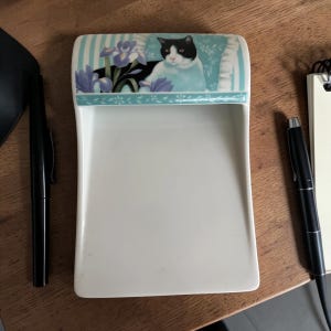 Puede incluir: Una bandeja de cerámica blanca con una parte superior decorativa que presenta un gato blanco y negro sobre un cojín azul con flores moradas. La bandeja tiene forma curva y está junto a dos bolígrafos negros.