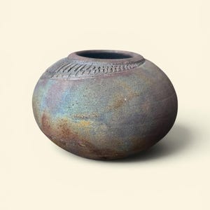 May include: A round, low-profile ceramic vase with a textured, dark matte finish. The surface displays iridescent hues of blue, purple, and copper. A decorative band of incised, repeating vertical lines encircles the upper shoulder area.