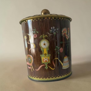 May include: A cylindrical metal storage canister with a dark brown wood grain finish and a gold-toned lid. The exterior features colorful illustrations of a grandfather clock, a rooster weather vane, a watering can, and floral motifs.