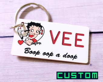 Peut inclure: Porte-clés rectangulaire blanc avec une image de dessin animé de Betty Boop et un chien dans un cœur rouge. Le porte-clés dit "VEE" en lettres rouges et "Boop Oop a Doop" en écriture cursive noire.