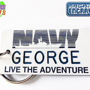 Puede incluir: Llavero de metal blanco con un diseño de barco de la marina azul y el texto "NAVY GEORGE LIVE THE ADVENTURE". El llavero está grabado a máquina.