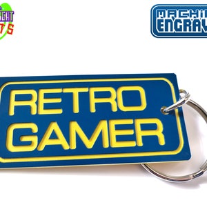 May include: Blue and yellow keychain with the text "RETRO GAMER" on it. The keychain is attached to a silver key ring. The text "MACHINE ENGRAVED" is visible in the top right corner of the image.