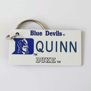 May include: White rectangular keychain with a silver ring. The keychain features the text "Blue Devils" in blue, the name "QUINN" in blue, and the word "DUKE" in black. A blue and white logo of a devil is on the left.
