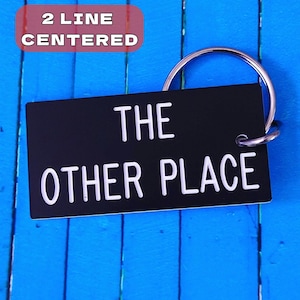 May include: A rectangular black keychain with white text that reads THE OTHER PLACE in two centered lines. The keychain is attached to a circular silver metal split ring. The design features clean, bold lettering on a solid black background.