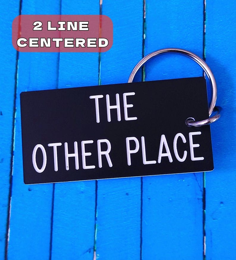 May include: A rectangular black keychain with white text that reads THE OTHER PLACE in two centered lines. The keychain is attached to a circular silver metal split ring. The design features clean, bold lettering on a solid black background.