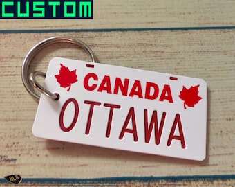 May include: White keychain with a red Canadian license plate design. The plate says "CANADA OTTAWA" with a red maple leaf on each side.
