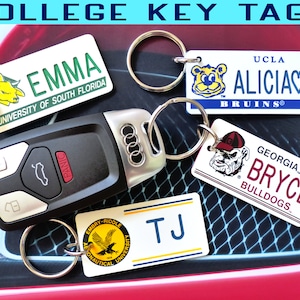 May include: A collection of college key tags with names and university logos. Key tags include "EMMA" from the University of South Florida, "ALICIA" from UCLA Bruins, "BRYCE" from Georgia Bulldogs, and "TJ" from Embry-Riddle Aeronautical University. An Audi car key is also present.