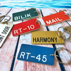 May include: A collection of rectangular metal keychains in various colors including white, dark green, red, gold, and blue. Each keychain features a metal split ring and engraved text reading FRONT DOOR, BILIK, MAIL, RT-10, HARMONY, and RT-45.