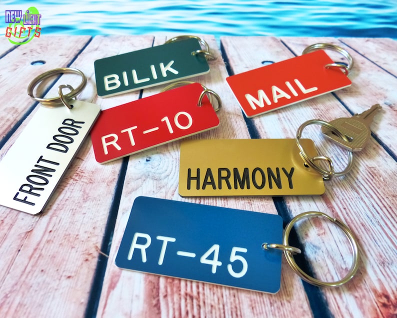 May include: A collection of rectangular metal keychains in various colors including white, dark green, red, gold, and blue. Each keychain features a metal split ring and engraved text reading FRONT DOOR, BILIK, MAIL, RT-10, HARMONY, and RT-45.