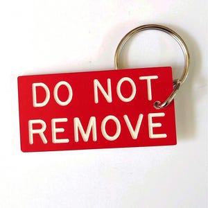 Könnte beinhalten: Ein rechteckiger roter Schlüsselanhänger mit dem weißen geprägten Text DO NOT REMOVE. Der Anhänger ist an einem runden silbernen Metallring befestigt. Das Design ist flach und kräftig, wobei der Text deutlich auf der roten Oberfläche zentriert ist.