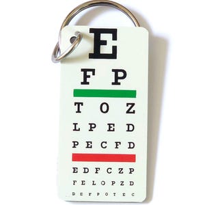 Puede incluir: Un llavero blanco con una tabla optométrica. La tabla presenta la letra "E" en la parte superior, seguida de filas de letras de tamaño decreciente. Hay una línea verde y una roja en la tabla.