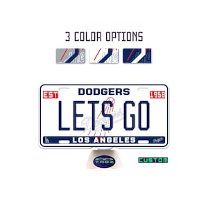 May include: A white license plate with the text "DODGERS LETS GO LOS ANGELES" in blue. The license plate has a red "EST 1958" box. The image also shows three color options and a "CUSTOM" text.