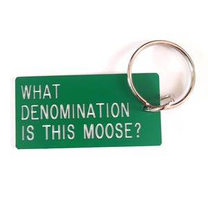 May include: A rectangular green keychain with a silver metal ring. The keychain features the white text WHAT DENOMINATION IS THIS MOOSE? engraved on the front surface. The design is simple and functional, made of a smooth, solid-colored material.