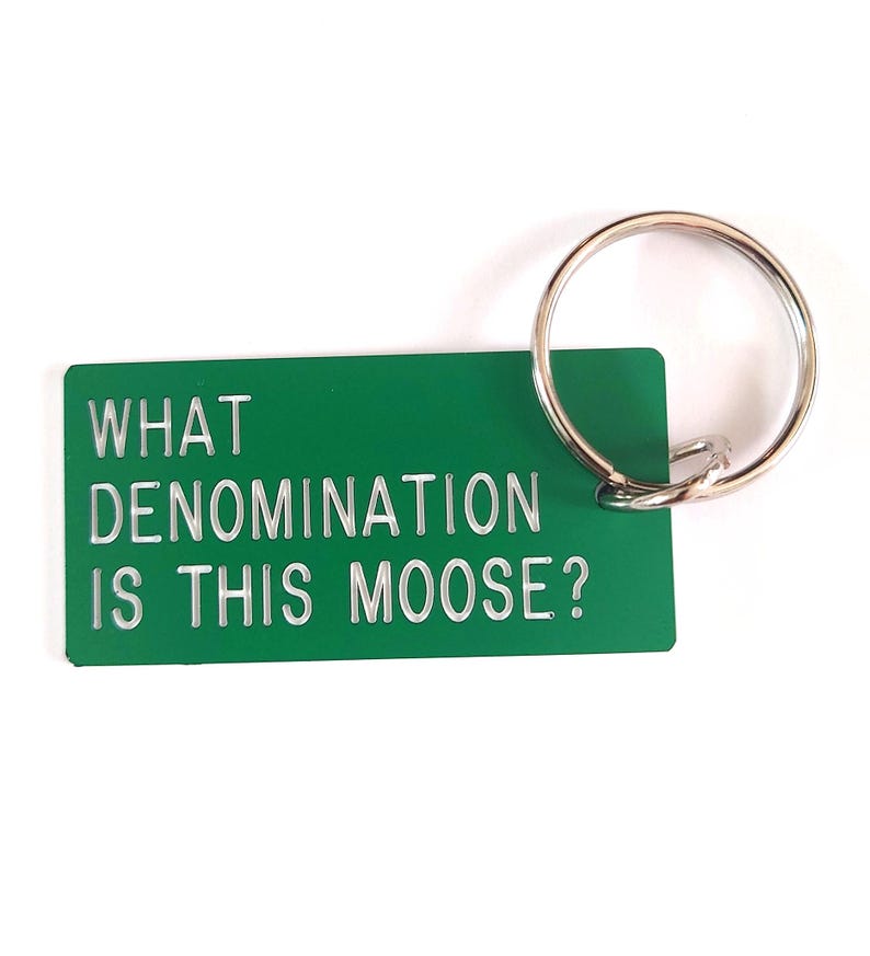 May include: A rectangular green keychain with a silver metal ring. The keychain features the white text WHAT DENOMINATION IS THIS MOOSE? engraved on the front surface. The design is simple and functional, made of a smooth, solid-colored material.
