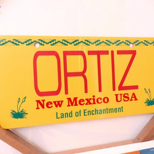 May include: Yellow metal license plate with a green and black border. The plate says "ORTIZ" in red letters, "New Mexico USA" in red letters, and "Land of Enchantment" in red letters.