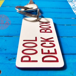 May include: A rectangular white keychain tag with rounded corners featuring the red engraved text POOL DECK BOX. The tag is attached to a silver metal split ring with a key. A red circular stamp with the text MADE IN THE USA is visible.