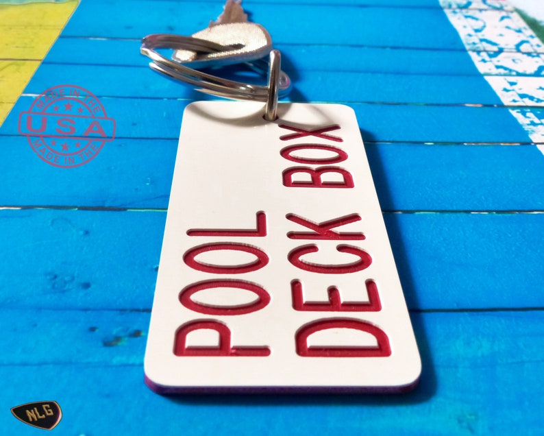 May include: A rectangular white keychain tag with rounded corners featuring the red engraved text POOL DECK BOX. The tag is attached to a silver metal split ring with a key. A red circular stamp with the text MADE IN THE USA is visible.