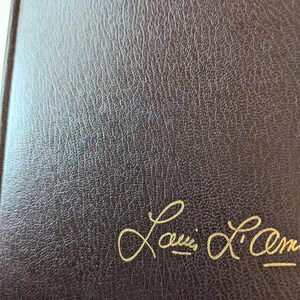 May include: A dark brown book cover with a textured, leather-like surface. The name Louis L'Amour is embossed in gold cursive script in the bottom right corner of the front cover. The material has a consistent grain pattern throughout the surface.