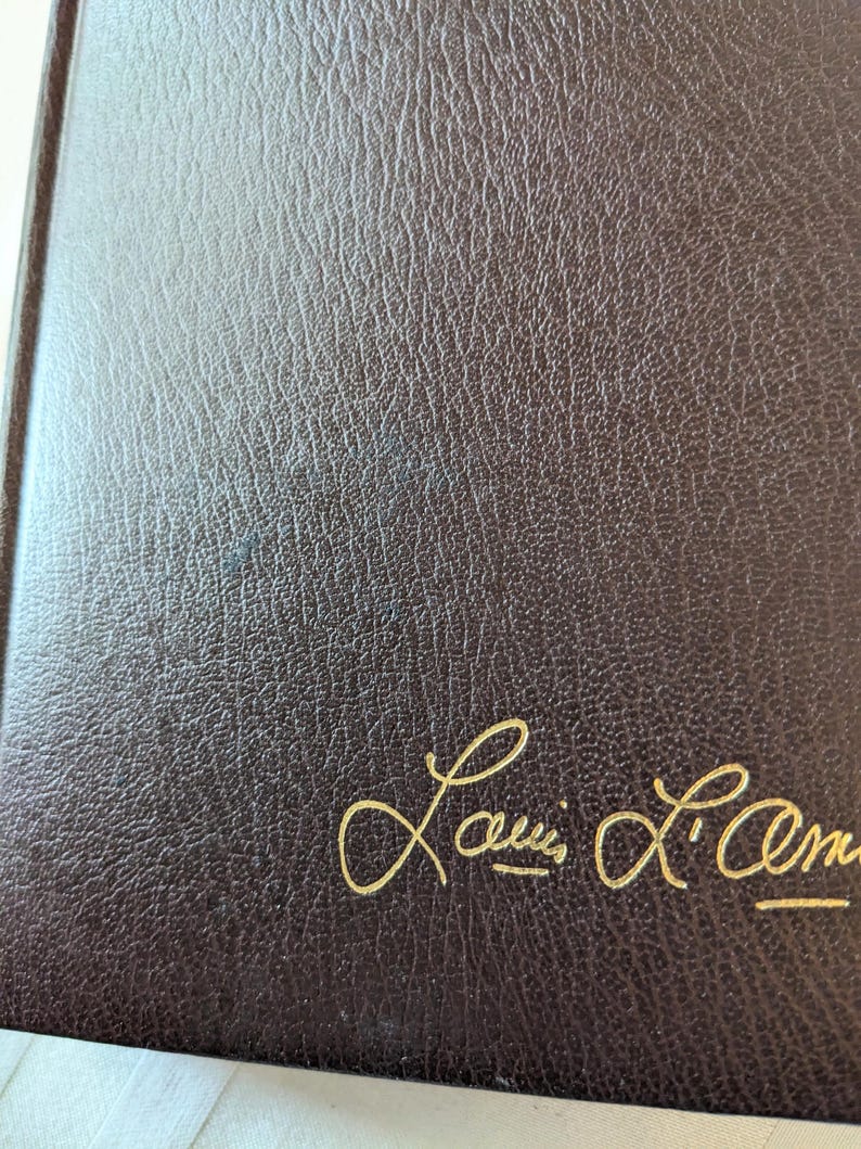 May include: A dark brown book cover with a textured, leather-like surface. The name Louis L'Amour is embossed in gold cursive script in the bottom right corner of the front cover. The material has a consistent grain pattern throughout the surface.