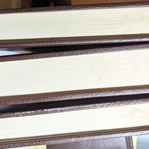 May include: Three hardcover books stacked horizontally, each featuring a dark brown textured cover. The pages are off-white with visible signs of age, including light yellowing and spotting along the edges of the paper blocks.