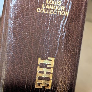 May include: A brown, textured, leather-like book spine featuring gold embossed lettering. The text reads THE LOUIS L'AMOUR COLLECTION at the top and THE WAL at the bottom. The surface has a distinct pebbled grain pattern throughout the material.