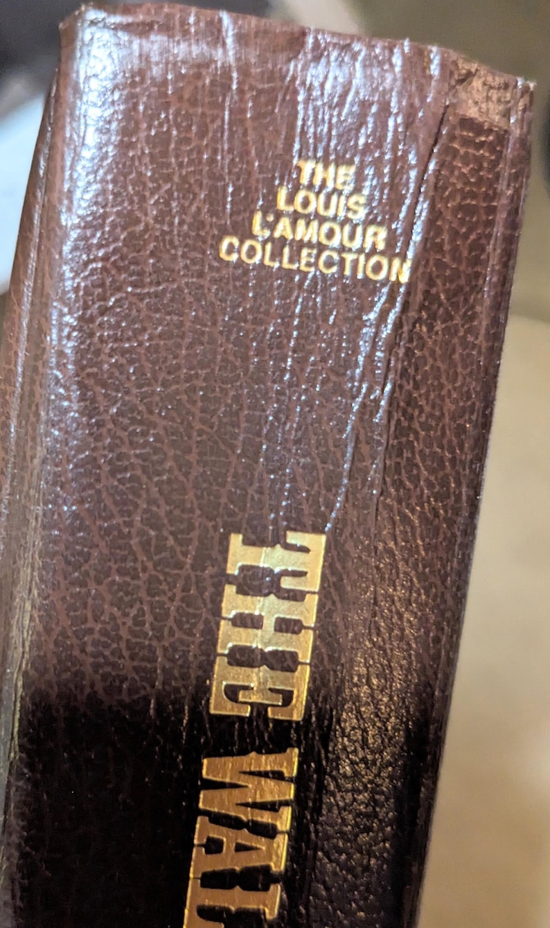May include: A brown, textured, leather-like book spine featuring gold embossed lettering. The text reads THE LOUIS L'AMOUR COLLECTION at the top and THE WAL at the bottom. The surface has a distinct pebbled grain pattern throughout the material.