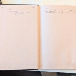 May include: An open hardcover book with light pink endpapers. The left page features handwritten text reading #7, 1982, and Marye Sanderson. The right page features handwritten text reading Sackett Series #1. A dark brown book cover is visible on the left.