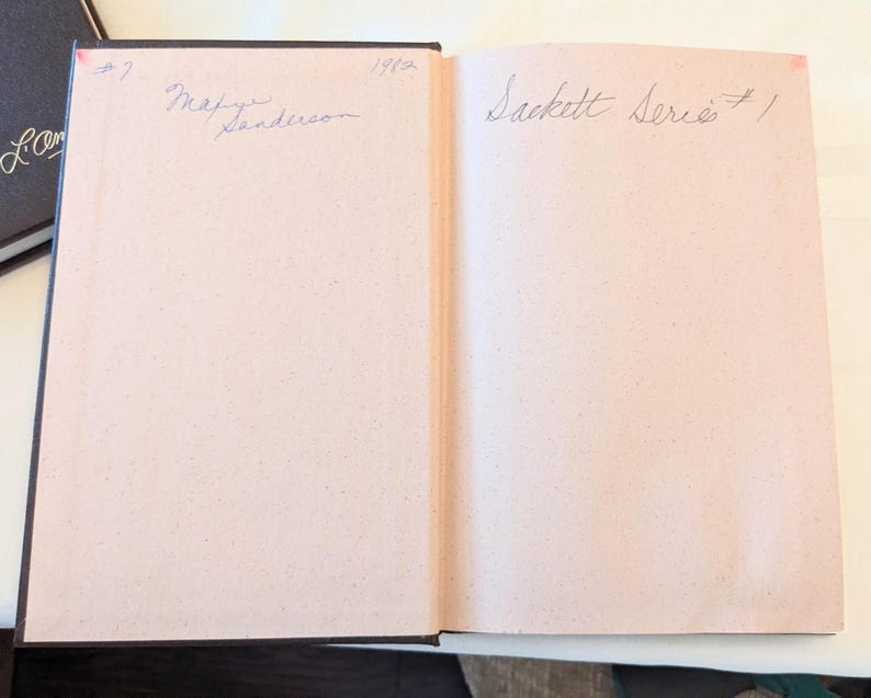 May include: An open hardcover book with light pink endpapers. The left page features handwritten text reading #7, 1982, and Marye Sanderson. The right page features handwritten text reading Sackett Series #1. A dark brown book cover is visible on the left.