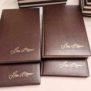 May include: A collection of hardcover books with dark brown, textured covers. Each book features the gold-embossed signature of Louis L'Amour on the front cover. The books are arranged in a stack, showcasing their uniform design and gold lettering.