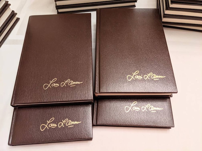 May include: A collection of hardcover books with dark brown, textured covers. Each book features the gold-embossed signature of Louis L'Amour on the front cover. The books are arranged in a stack, showcasing their uniform design and gold lettering.