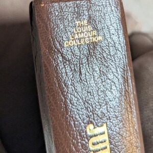 May include: A brown book spine with a textured surface featuring gold embossed text that reads THE LOUIS LAMOUR COLLECTION at the top and JUBAL at the bottom. The book has a vertical orientation and a dark brown finish with visible grain patterns.