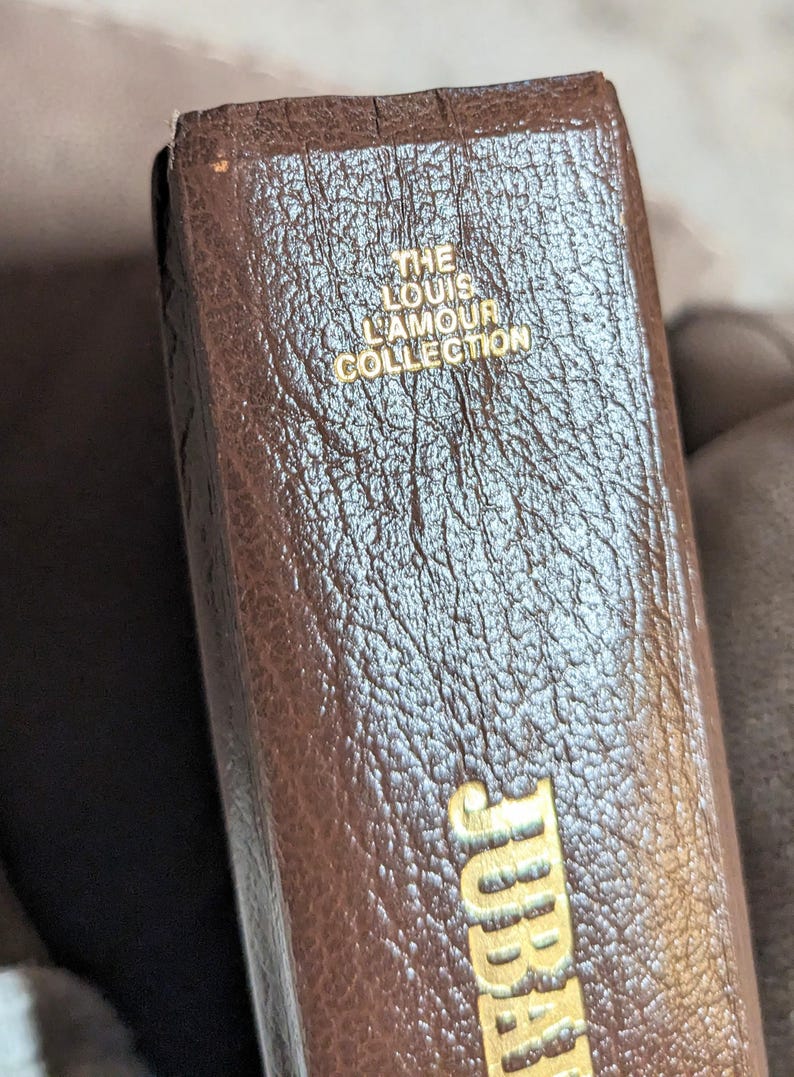 May include: A brown book spine with a textured surface featuring gold embossed text that reads THE LOUIS LAMOUR COLLECTION at the top and JUBAL at the bottom. The book has a vertical orientation and a dark brown finish with visible grain patterns.