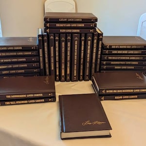 May include: A collection of Louis L'Amour books with dark brown covers and gold lettering. The set includes multiple volumes arranged in stacks and standing upright, featuring titles such as Catlow, The Empty Land, Flint, and The Daybreakers.