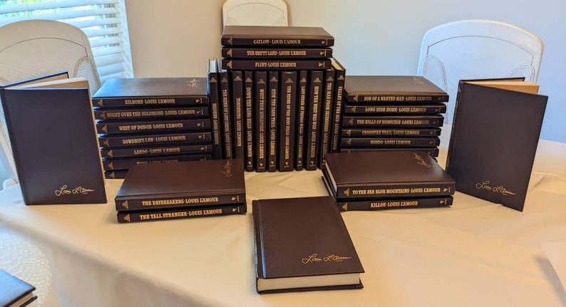 May include: A collection of Louis L'Amour books with dark brown covers and gold lettering. The set includes multiple volumes arranged in stacks and standing upright, featuring titles such as Catlow, The Empty Land, Flint, and The Daybreakers.